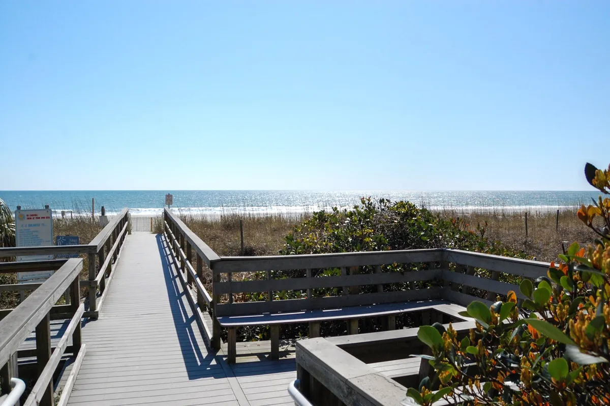 A scenic boardwalk leading towards the ocean, bordered by dune grass and open sky. The view showcases easy access to the beach and opportunities for sunrise walks or leisurely exploration.