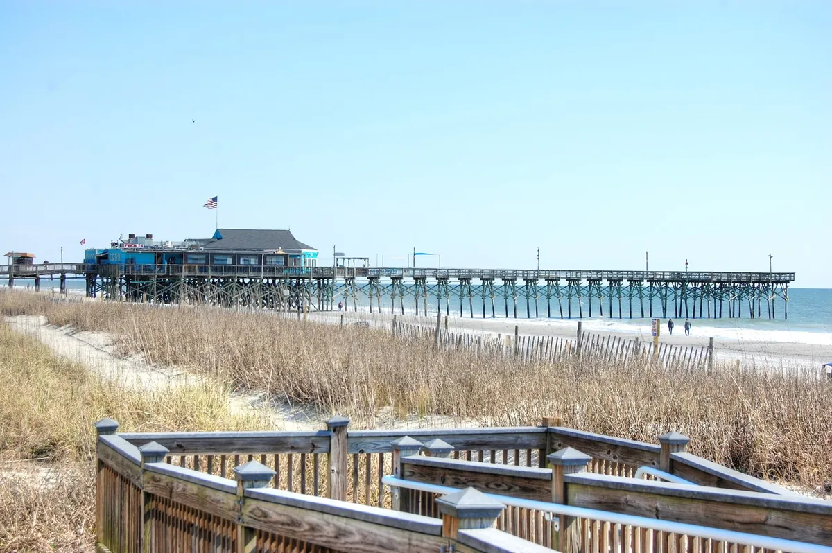 A tranquil beach scene with a long pier stretching over the water, soft sand, dune grass, and a bright blue sky. The image invites viewers to imagine fishing off the pier, building sandcastles, or simply soaking up the sun beside the ocean.