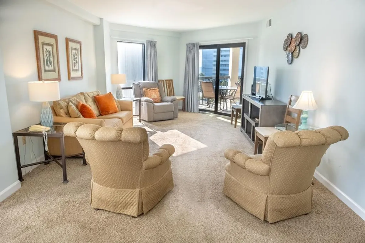 The image features a comfortable seating area with a sofa adorned with orange cushions, two armchairs, a television, and a sliding glass door that hints at accessibility and openness. Decorative wall art adds a touch of hospitality, while the space feels ready for relaxation or socializing.