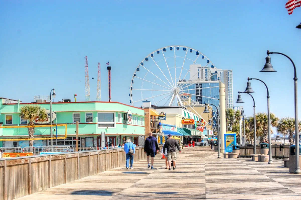 A lively boardwalk scene featuring a ferris wheel, palm trees, shops, and people strolling and enjoying amusement rides. The image captures the festive atmosphere of a classic coastal destination, perfect for shopping, riding attractions, and watching sunsets.