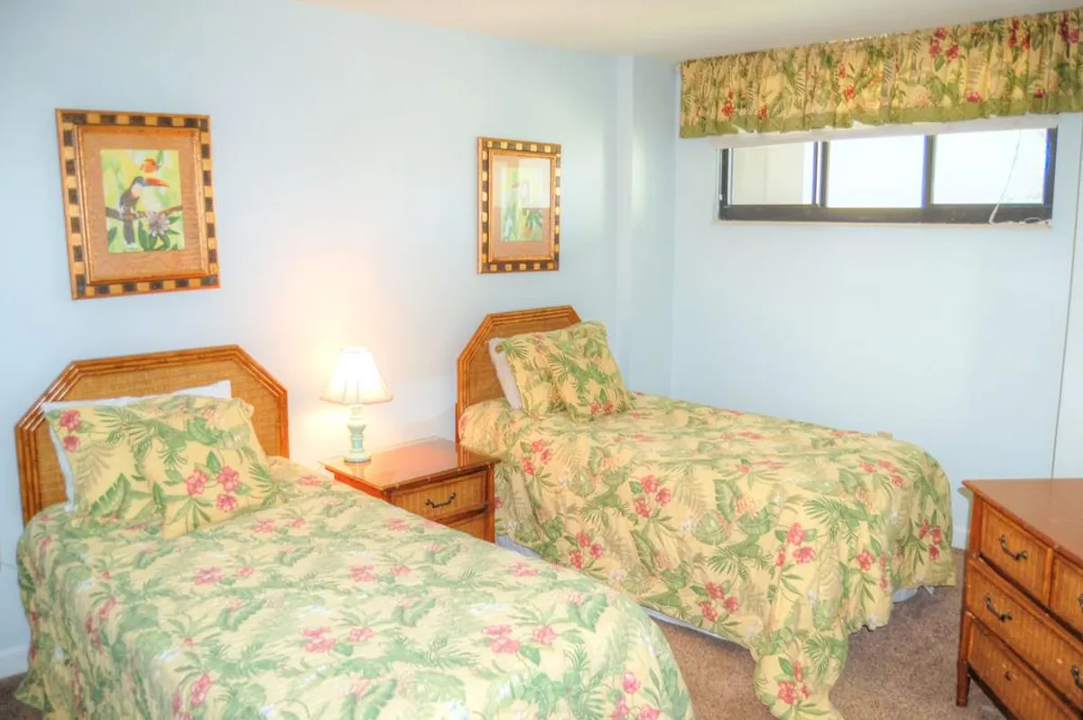 A welcoming bedroom with twin beds dressed in floral bedding, a nightstand with a lamp, and a window letting in natural light. Decorative wall art and soft hues create a tranquil, family-friendly environment.