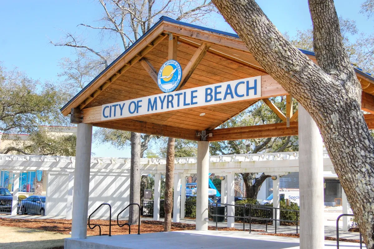 A welcoming wooden structure with 'City of Myrtle Beach' signage, surrounded by trees. The scene hints at community activities, events, and the vibrant local environment beyond the beach.