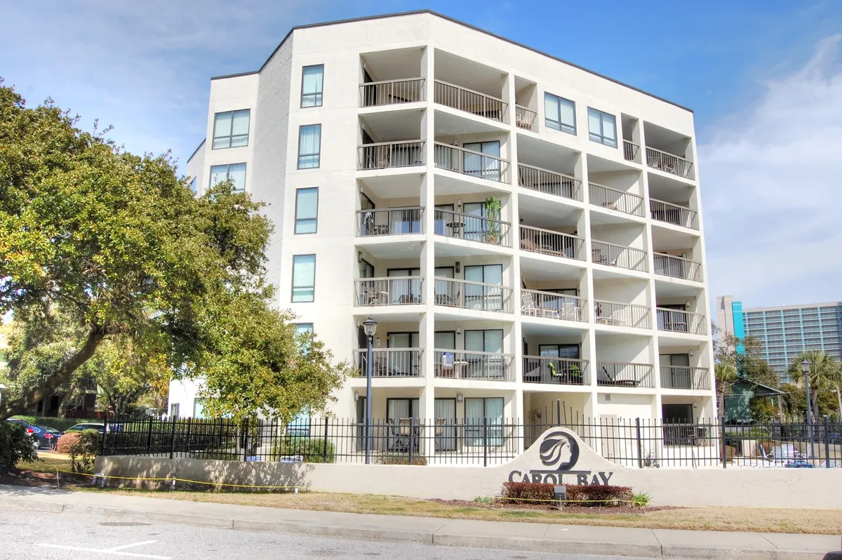 The image highlights the building's attractive balconies, entrance sign, and lush surrounding greenery. The balconies suggest inviting spaces for groups to gather and enjoy sunsets or morning coffee together. The communal feel and exterior context connect directly with group vacations and shared experiences.