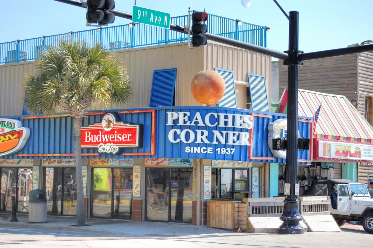 The image features the iconic Peaches Corner signage, Budweiser logo, a large orange ball decoration, and palm trees, set against an energetic street scene. The lively atmosphere reflects the vibrant dining and nightlife options close to Carol Bay.