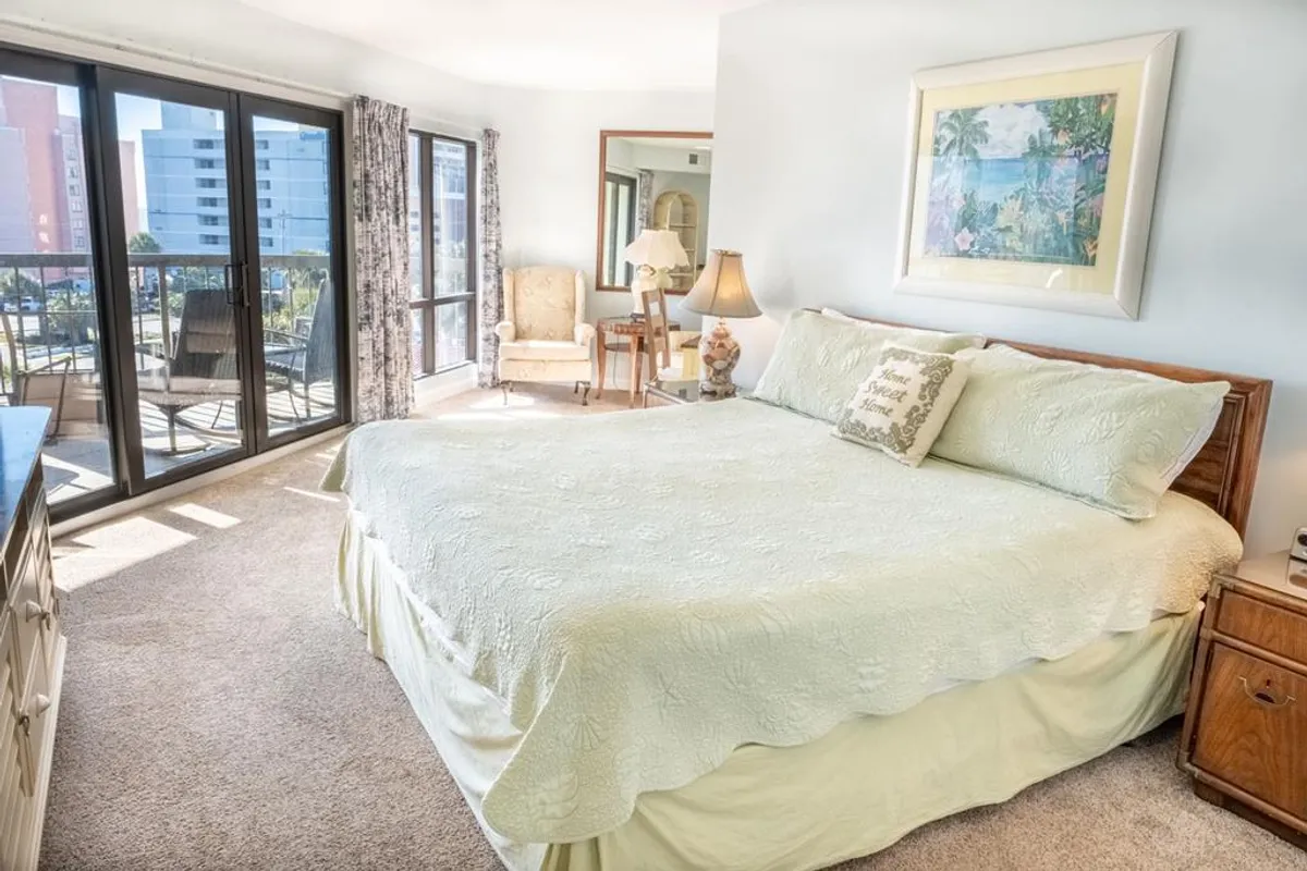 A spacious bedroom featuring a plush bed positioned to capture sunrise views through large windows. The room is tastefully decorated with an armchair, lamp, and framed artwork, creating a comforting and serene retreat.