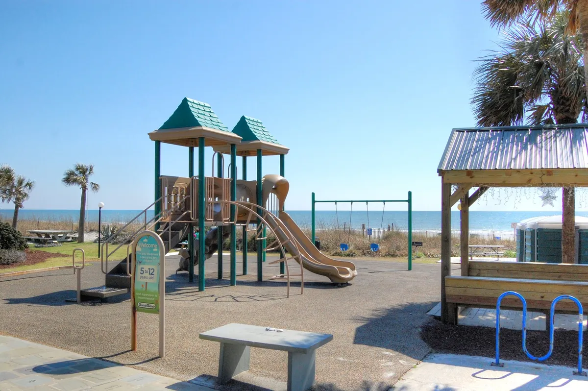 This vibrant exterior image showcases a playground with swings and other play equipment, families and children enjoying the area, and a beach view framed by palm trees, with a picnic area nearby. The scene is filled with people making the most of a sunny day, embodying active family fun and relaxation by the ocean.