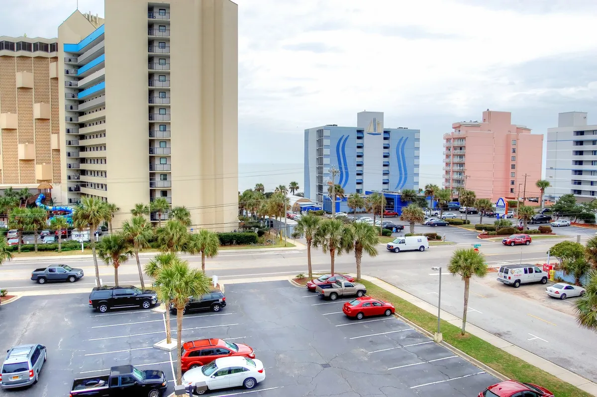 This exterior image showcases palm trees, hotel buildings, and a parking lot, all bathed in vacation-ready light. The scene hints at easy access to both accommodations and the lively local Myrtle Beach area, suggesting convenience for exploring attractions and participating in complimentary activities.