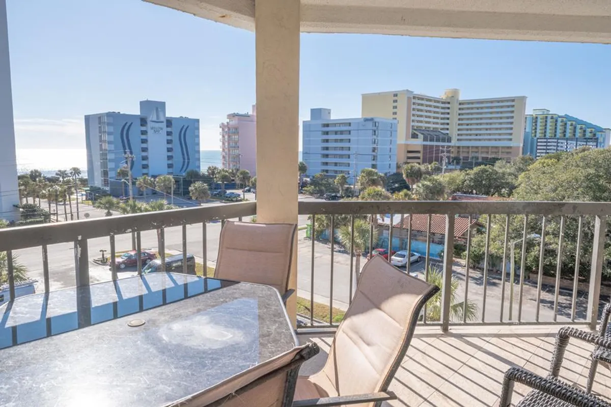 This image captures a tranquil balcony scene with comfortable seating, offering a sweeping view over the pool area, the ocean, and surrounding buildings. The serene setting is ideal for relaxing, enjoying a meal outdoors, or watching sunrise and sunset with the ocean breeze.